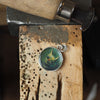 Oil-painted silver pendant resting on a jeweller’s bench, shown during the making process.