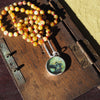 Custom painted pendant on a beaded necklace, displayed on a wooden box in natural light.