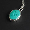Turquoise intaglio dog necklace at an angle on black, the depth of the carving clear across the stone and the silver bezel catching the light